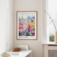 a framed poster of a colorful floral scene hanging on a wall, with a window and a small table with books and a vase in the foreground.