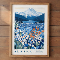The image is a framed poster depicting a scenic landscape of Alaska, featuring a mountain range, a lake, and a field of wildflowers.