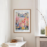 a framed poster of a colorful floral scene hanging on a wall, with a white table and a vase of flowers in the foreground.