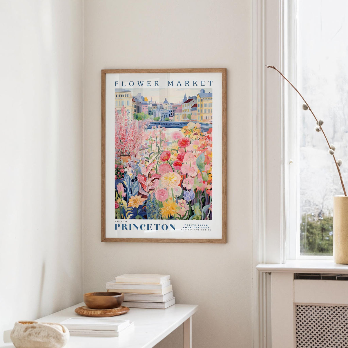 a framed poster of a colorful floral scene hanging on a wall, with a white table and a vase of flowers in the foreground.