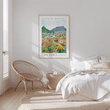 a bedroom with a bed, a woven chair, and a framed poster of a flower market in Cape Town hanging on the wall.