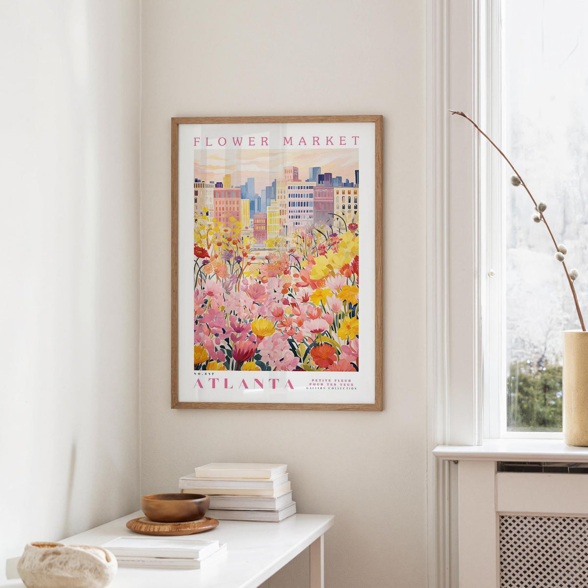 a framed poster of a colorful floral scene hanging on a wall, with a window and a small table with books and a vase in the foreground.