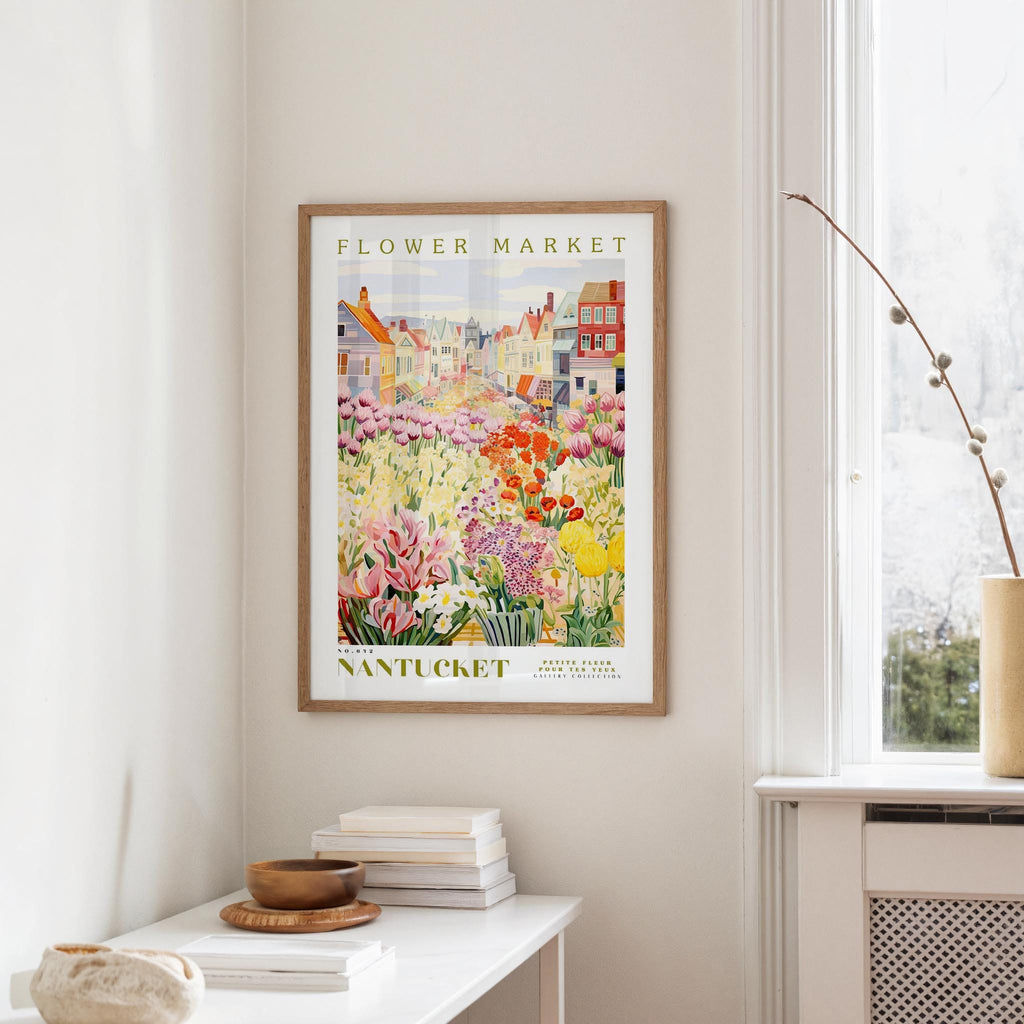a framed poster of a colorful garden scene hanging on a wall, with a white table and a vase of flowers in the foreground.
