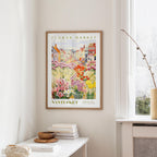 a framed poster of a colorful garden scene hanging on a wall, with a white table and a vase of flowers in the foreground.