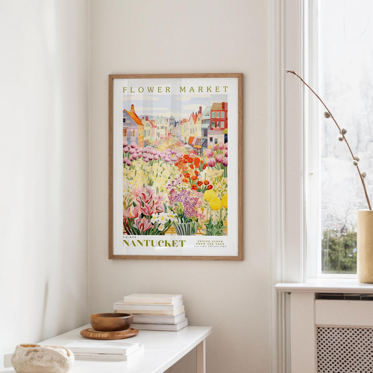 a framed poster of a colorful garden scene hanging on a wall, with a white table and a vase of flowers in the foreground.