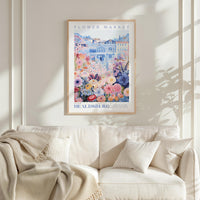 a framed poster of a colorful floral scene hanging on a wall above a white couch with a blanket draped over it.