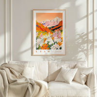 a framed poster of a mountainous landscape with a vibrant, colorful scene of flowers and foliage. The poster is hung on a wall above a white couch, which is adorned with a blanket and pillows.