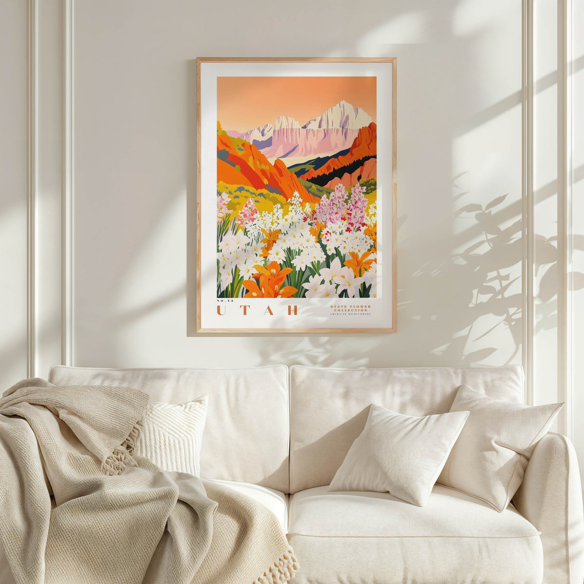 a framed poster of a mountainous landscape with a vibrant, colorful scene of flowers and foliage. The poster is hung on a wall above a white couch, which is adorned with a blanket and pillows.