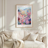 a framed poster of a colorful floral scene hanging on a wall above a white couch with a blanket draped over it.