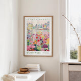 a framed poster of a colorful floral scene hanging on a wall, with a window and a small table with books and a vase in the foreground.