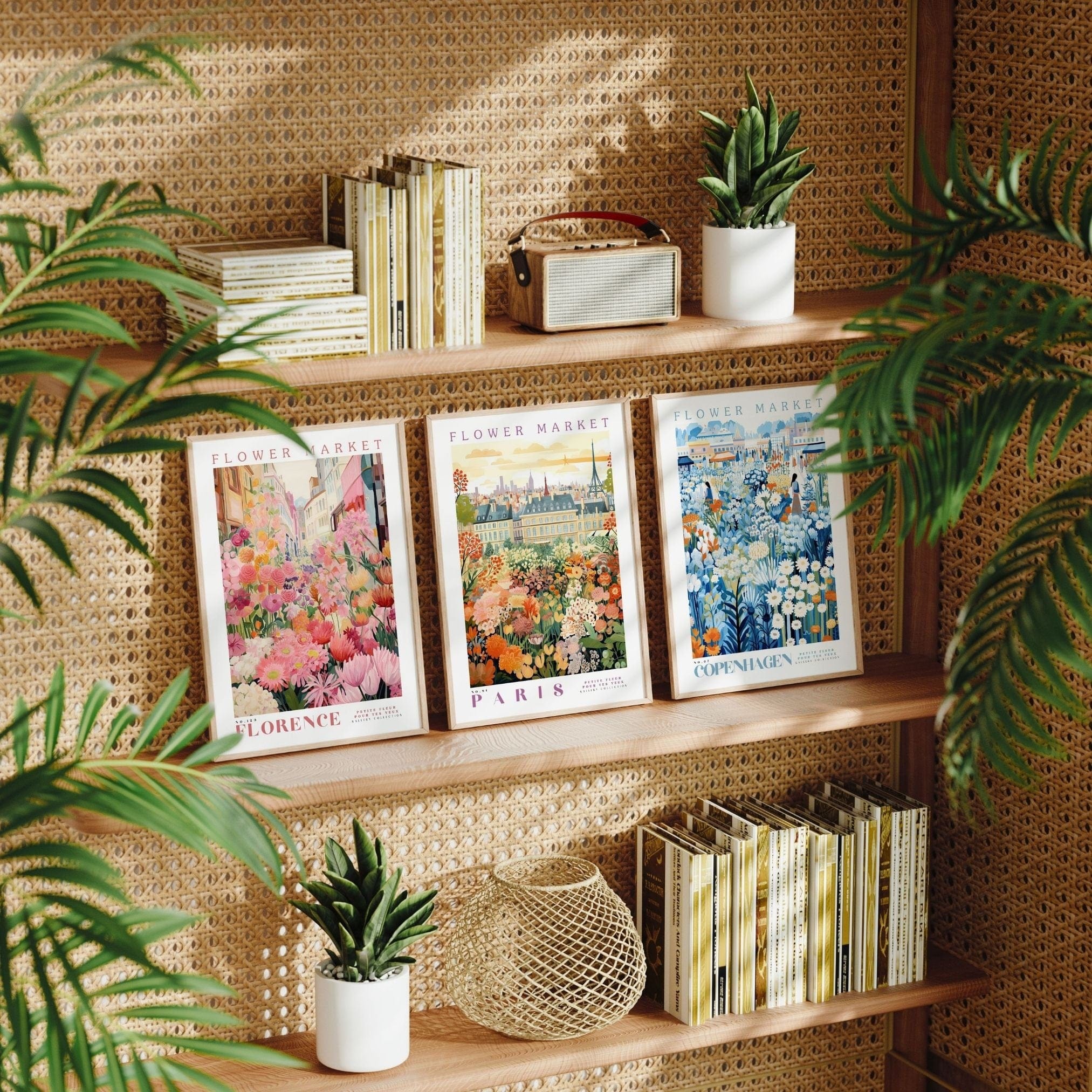 Three framed flower market posters displayed on wooden shelves with a woven rattan backdrop: Florence with vibrant pink and orange blooms, Paris featuring lush green and orange florals, and Copenhagen with blue and white flowers. The scene includes potted plants, vintage books, and a decorative woven basket, surrounded by green palm leaves, creating a cozy and stylish natural aesthetic.